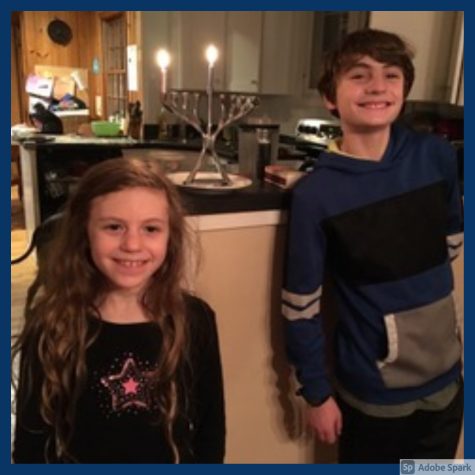 A photo of Raphael Gold standing to the right of the frame in a blue sweatshirt next to a menorah with 2 candles lit and Dalia Fermaglich standing next to him on his left 