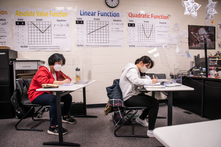 Junhwan Ahn (9) and Nijiko Nomura (9) writing on their notebooks in their seat. Posters of "Absolute Value function," "Linear Function" and "Quadratic Function" are hanged on the wall. 