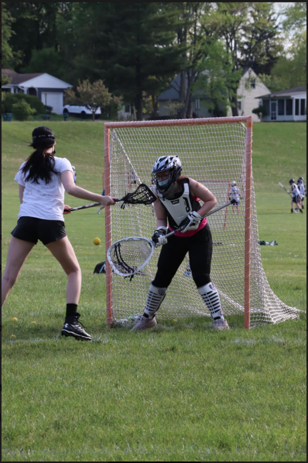 a girl holding a lacrosse stick runs up to a goal with another girl acting as goalie.