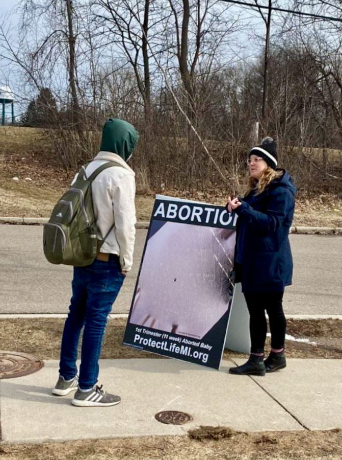 Woman with blond hair and blue coat stands next to folded sign saying ABORTION on the top with a blurred image and Protectlifemi.org written under the photo. A student in a green hoodie, white jacket, and jeans stands talking to her.